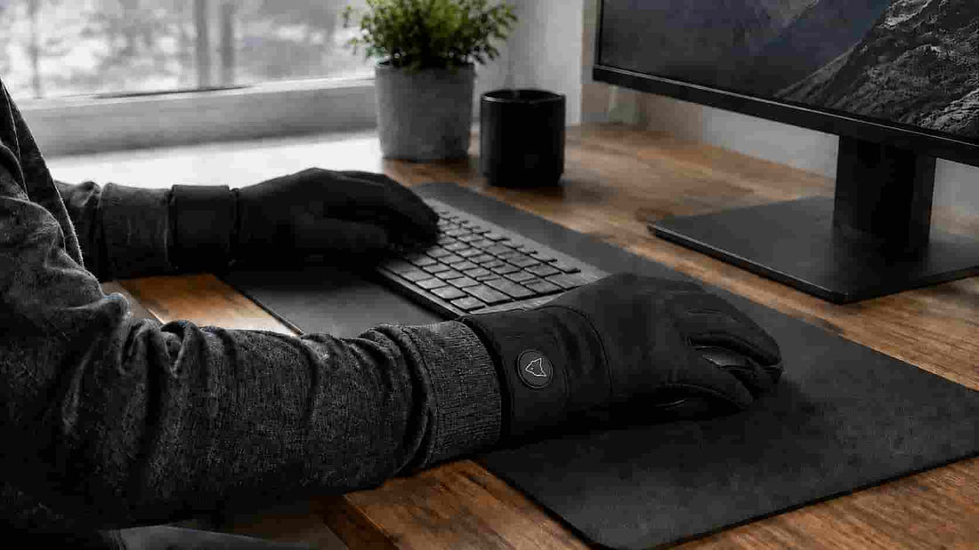 Person wearing black heated gloves with a visible wrist heat control while working at a computer in a cool home office