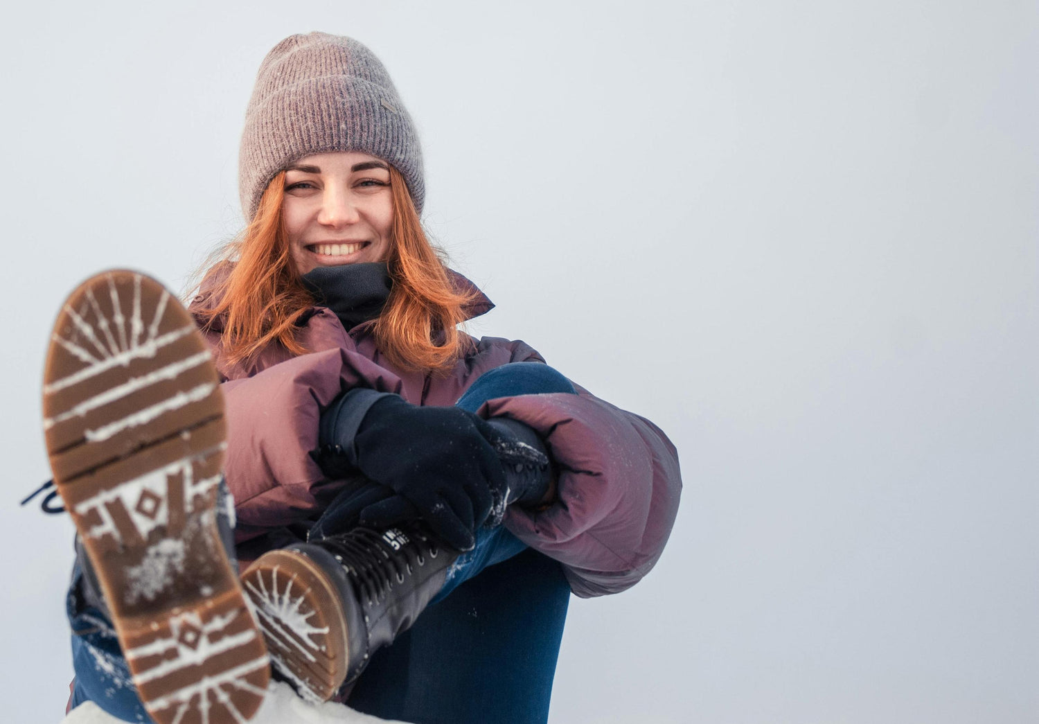 A smiling woman sitting in the snow wearing a winter puffer jacket, beanie, and Arcfomor slim-fit rechargeable heated gloves for women.