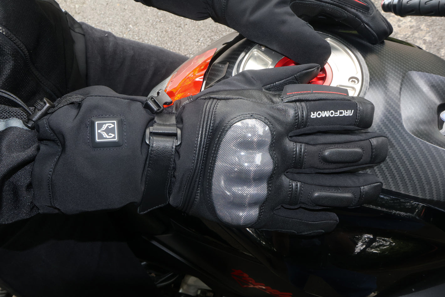 Close-up of the carbon fiber knuckle shell on Basalt heated motorcycle gloves beside a motorcycle fuel tank
