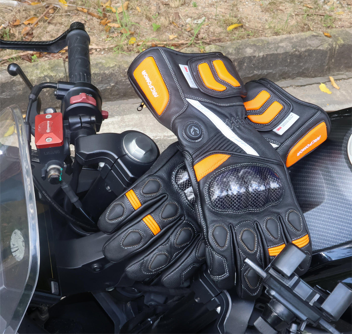 Both black and orange Tempest heated motorcycle gloves shown beside a motorcycle handlebar