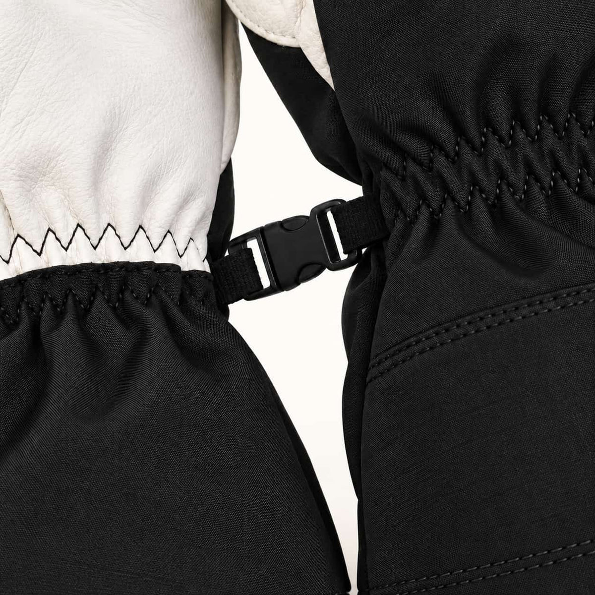 Black pairing clip joining the elastic wrists of two Yellowstone gloves, beside white leather and black woven fabric.